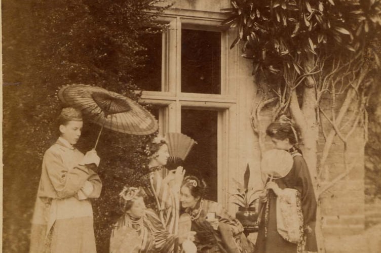 Group of women dressed in Japanese costume, outside a window of a large house. Two hold fans and one a parasol.