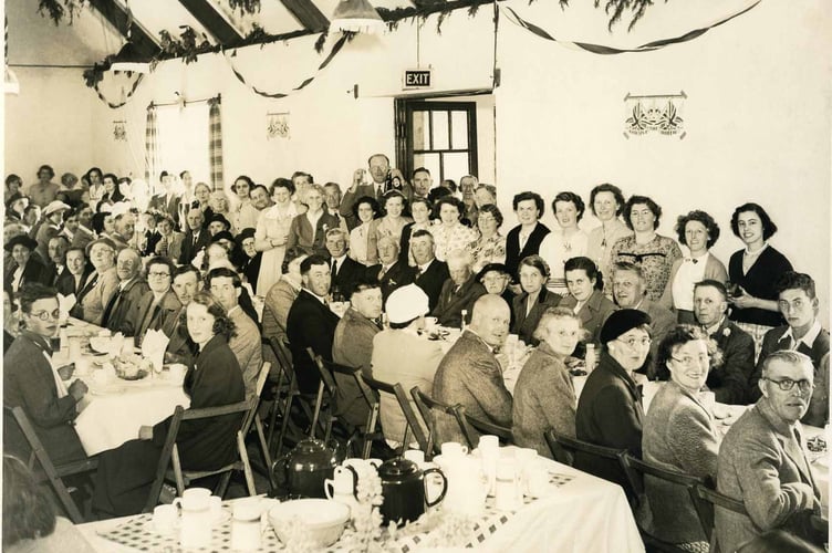 Photograph of a group of people in the village hall in Chillington. Includes H.Lee the postman. As the poster on the wall reads "God Save the Queen"  it is possibly 1953. Includes Beatrice Lee, Henry Lee  and daughter Mary Lee. On the table seated right hand side, 5-3 from right.